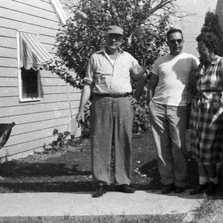 Three people stand near a vintage car and house.