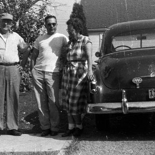Three people stand near a vintage car and house.