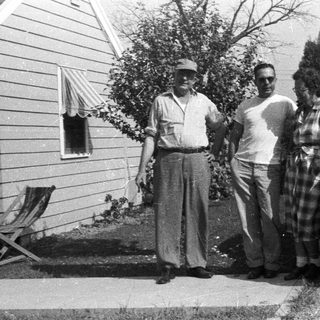 Three people stand near a vintage car and house.