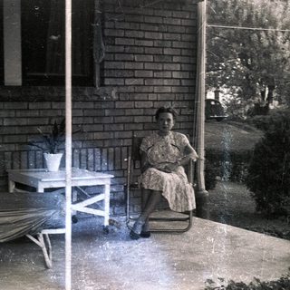 Woman sits on porch with furniture and plants.