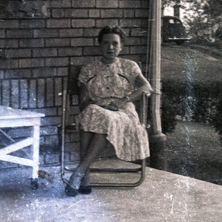 Woman sits on porch with furniture and plants.