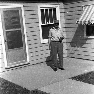 Man stands outside a house, partially shaded by an awning.