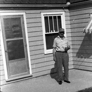 Man stands outside a house, partially shaded by an awning.
