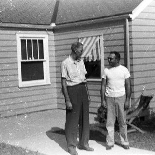 Two men stand outside a modest, older house.