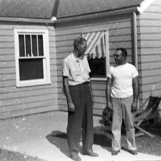 Two men stand outside a modest, older house.