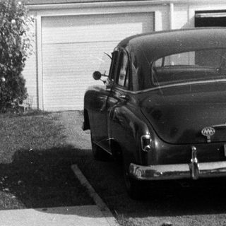 Woman stands on sidewalk near vintage car and house.