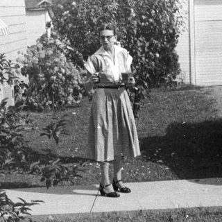Woman stands on sidewalk near vintage car and house.