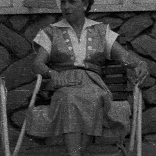 Woman sits on a chair outside a stone building.