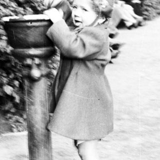 Here's a brief summary of the image: Child stands at a vintage fountain, reaching for water.