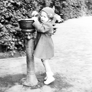 Here's a brief summary of the image: Child stands at a vintage fountain, reaching for water.