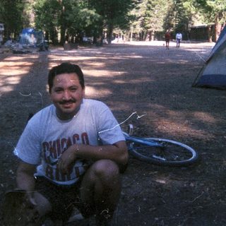 Man sits near tent in a wooded campsite.