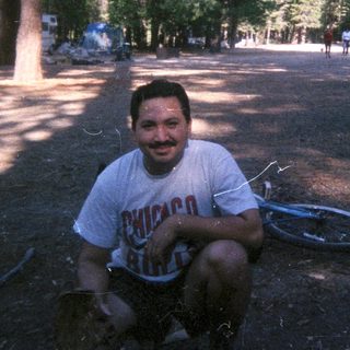 Man sits near tent in a wooded campsite.
