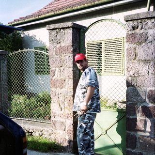 Man in camouflage stands near a stone building and car.