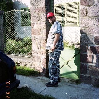 Man in camouflage stands near a stone building and car.