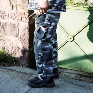 Man in camouflage stands near a stone building and car.