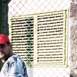Man in camouflage stands near a stone building and car.