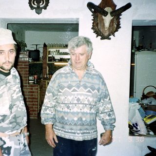 Two men stand indoors, with a deer head mounted on the wall.