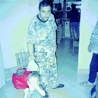 Man in camo pants stands near a child in a kitchen.