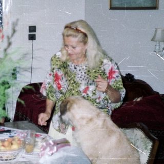 Woman and dog sit near table with fruit and flowers.