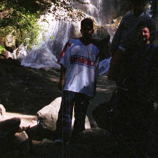 People stand near a waterfall in a lush, rocky area.