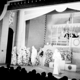 Stage scene with performers and a ship backdrop.