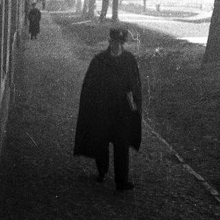 Man in a cloak walks along a tree-lined path.