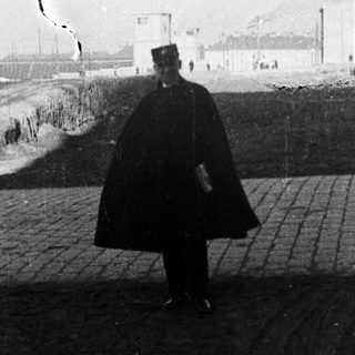 Man in cloak stands before a building complex.