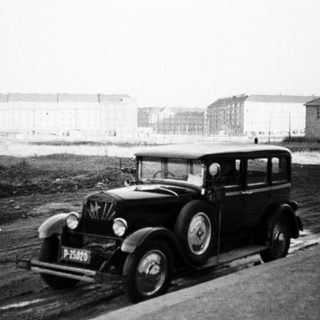 Vintage car travels on a road with buildings in background.