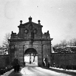 Monochrome image shows a gate and snowy, walled road.