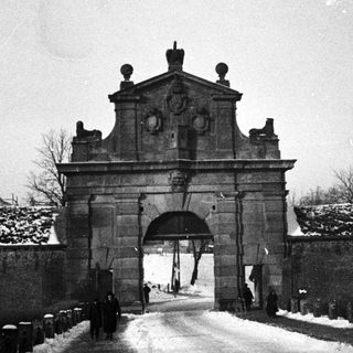 Monochrome image shows a gate and snowy, walled road.