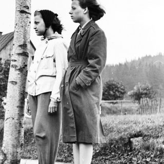 Two young women stand near a tree in a grassy area.