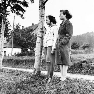 Two young women stand near a tree in a grassy area.