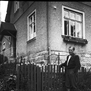 Man stands near a house with a picket fence.