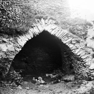 Ruined stone archway, overgrown with vegetation, sits in landscape.