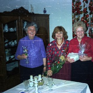 Three women stand smiling, holding gifts in a dining room.