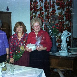 Three women stand smiling, holding gifts in a dining room.