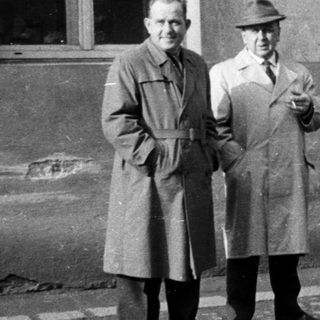 Five men stand outside a building, dressed in coats.