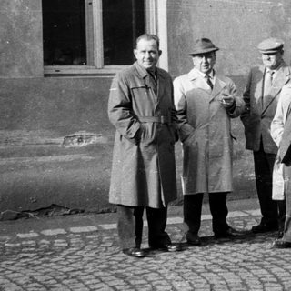 Five men stand outside a building, dressed in coats.