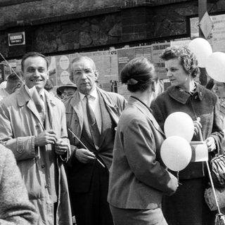 People holding balloons stand near a poster-covered wall.