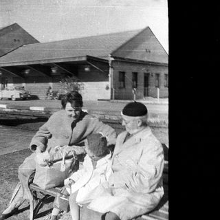 Here's a brief summary of the image: Soldier and family sit on a bench, likely at a station.
