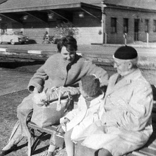 Here's a brief summary of the image: Soldier and family sit on a bench, likely at a station.