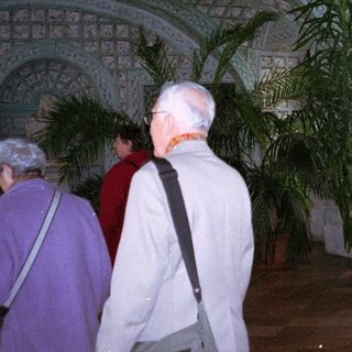 People tour an ornate, arched hall with potted plants.