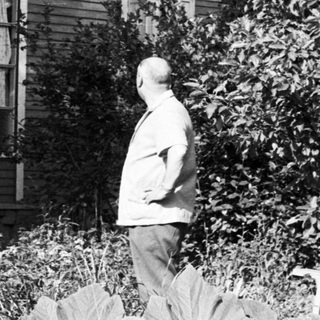 Man stands near house, surrounded by trees and foliage.