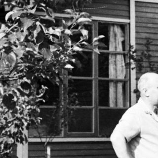 Man stands near a house in a wooded, overgrown area.