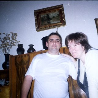 Two people pose indoors near a dark wood cabinet.