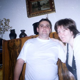 Two people pose indoors near a dark wood cabinet.