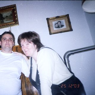 Two people pose indoors near a dark wood cabinet.