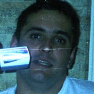 Man seated, holding a video camera indoors, dimly lit.
