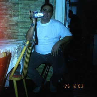 Man seated, holding a video camera indoors, dimly lit.
