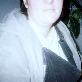 Person wearing a hooded sweatshirt and overalls, plants in background.